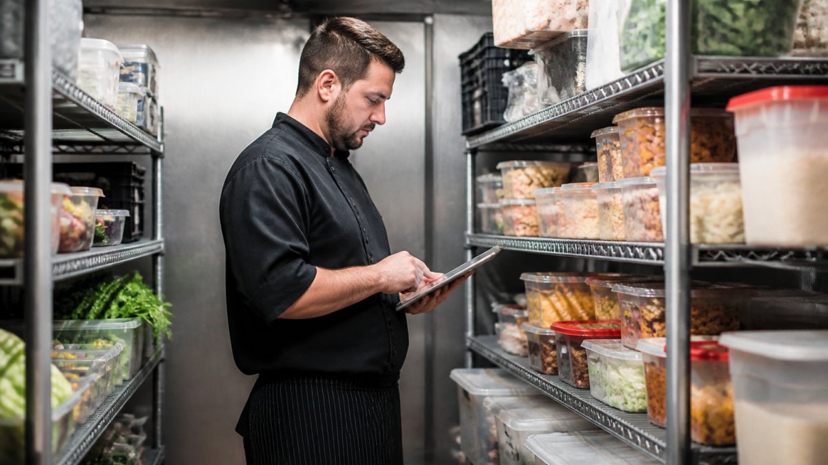 Restaurant manager reviewing inventory levels on tablet while standing in organized walk-in cooler with labeled ingredient containers and organized shelves