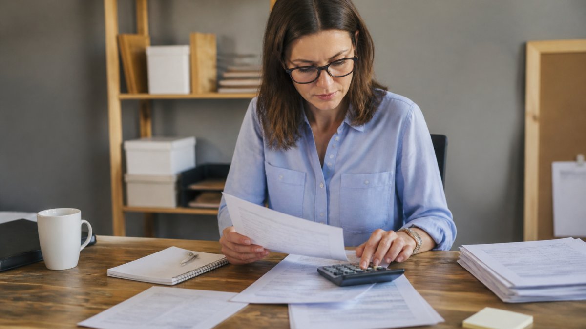 Small business owner at desk with laptop, bank statements, and calculator, reviewing financial records with focused concentration in natural office lighting